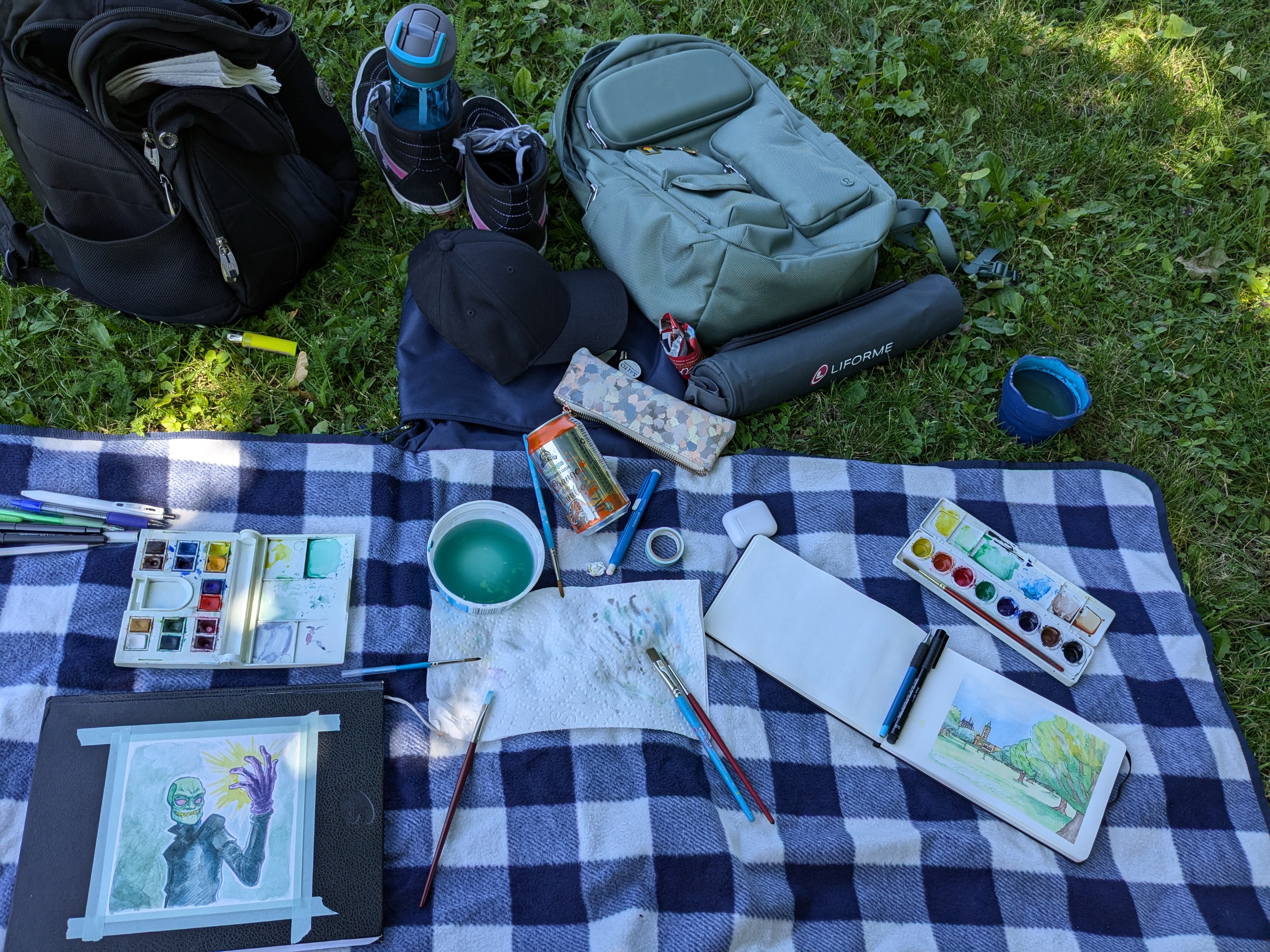 A photo of watercolour materials spread out on a picnic blanket with two different paintings, as well as two backpacks, a pair of shoes, and other miscellany